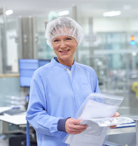 Hymyilevä nainen laboratoriovarustus päällään pitelee papereita käsissään.